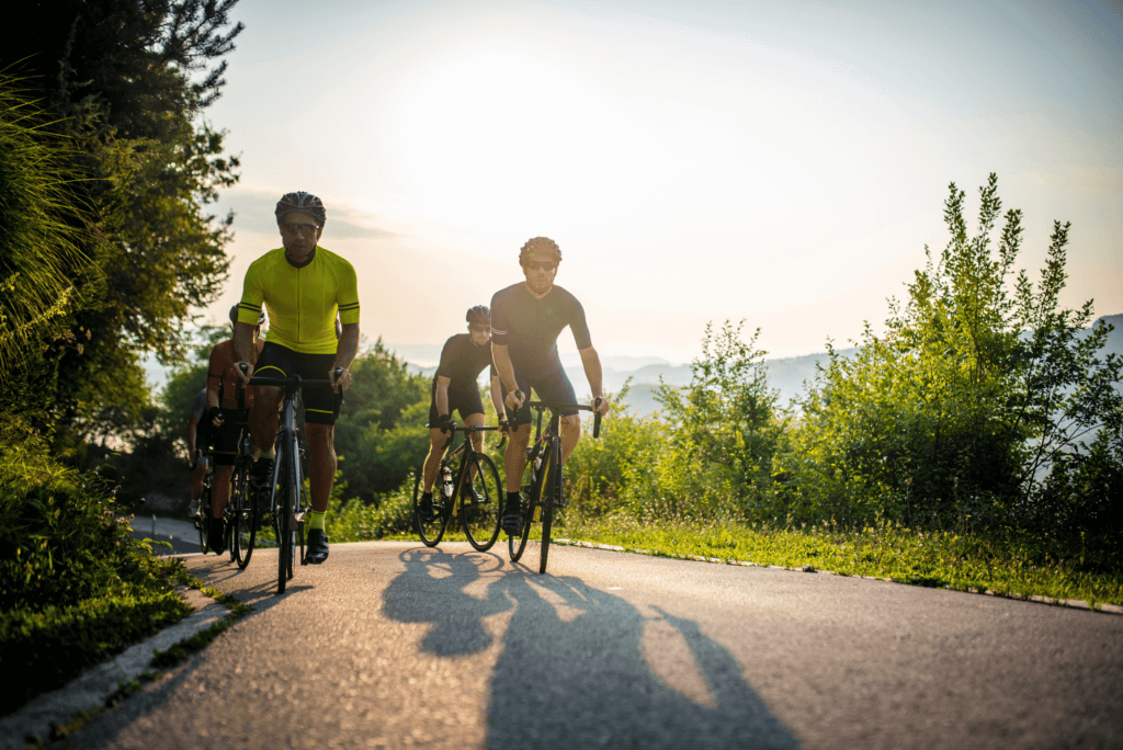 three male cyclist going up a hill with trees and the sun behind them