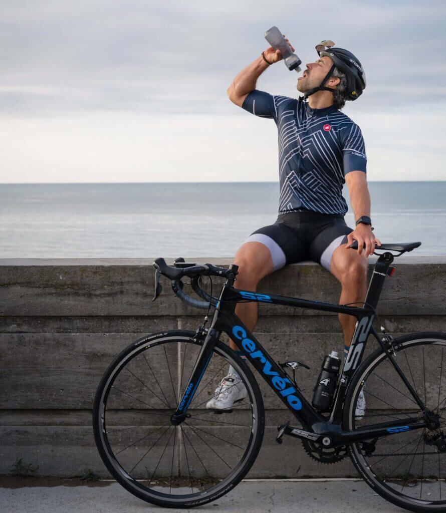 a cyclist sitting on a wall drinking from his bottle, with his road bike in front of him