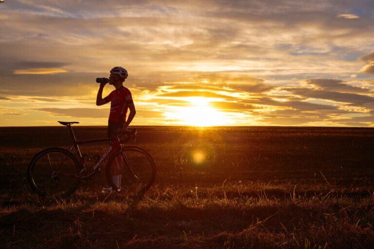 a male cyclist standing beside his bike, drinking from a bottle with the sun setting behind him
