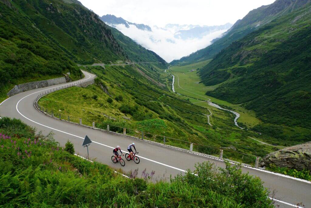 two cyclists cycling up hill with mountains behind them