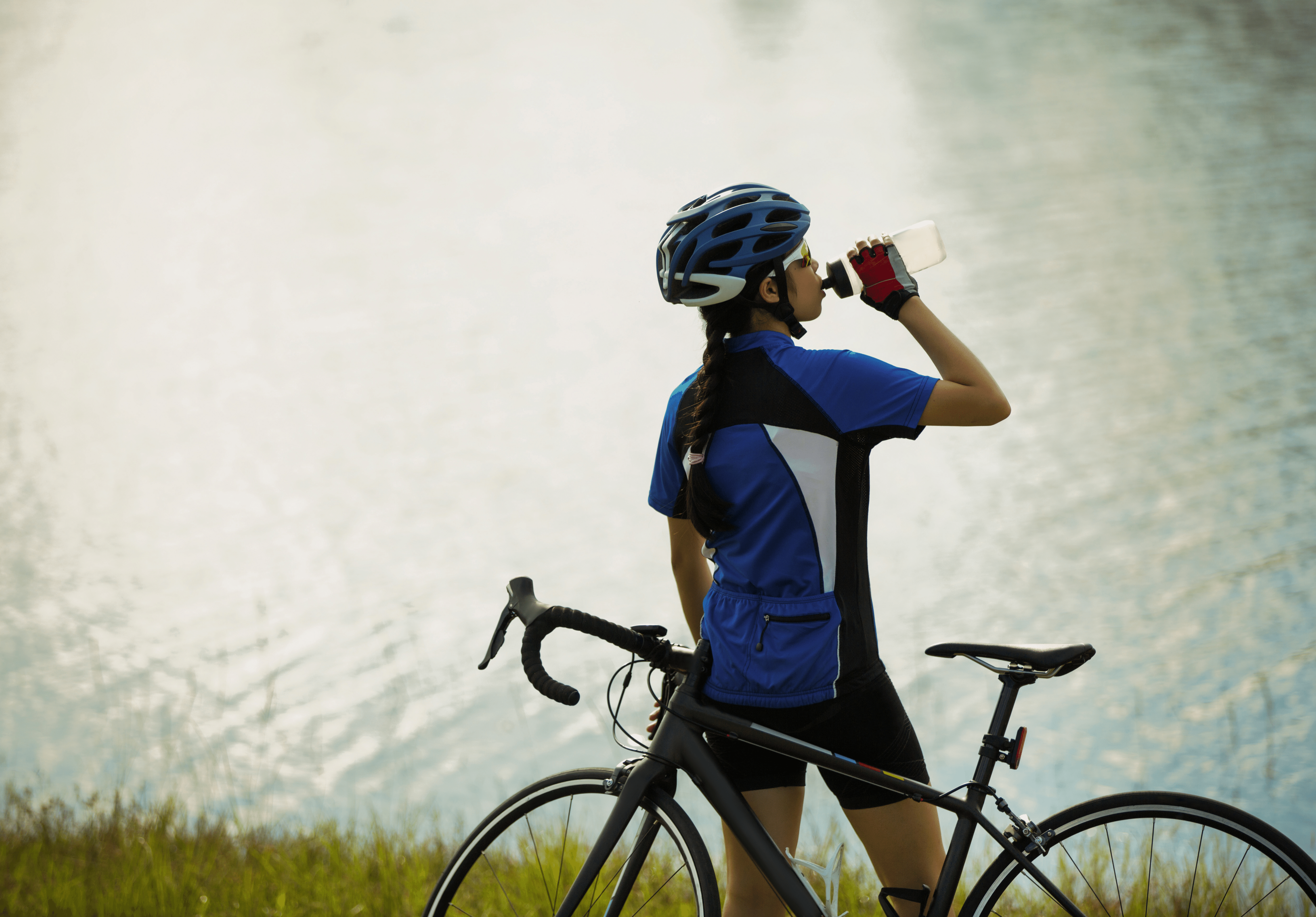 a woman cyclist standing by her bike, looking out on a lake, drinking from a bottle