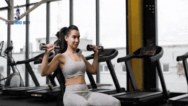 a woman in a gym, sitting down doing overhead presses with dumbells