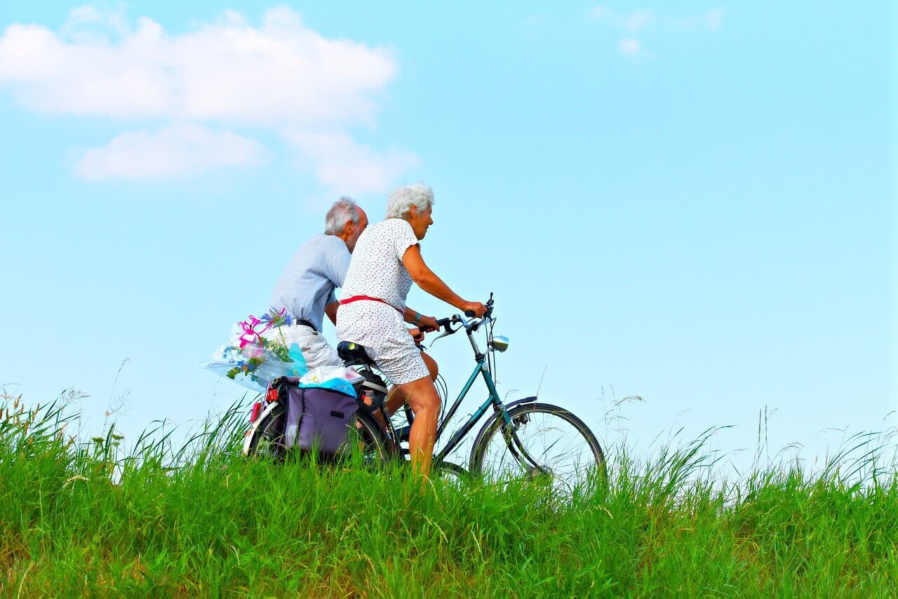 an older man and woman cycling together beside grass fields, with groceries in their bags