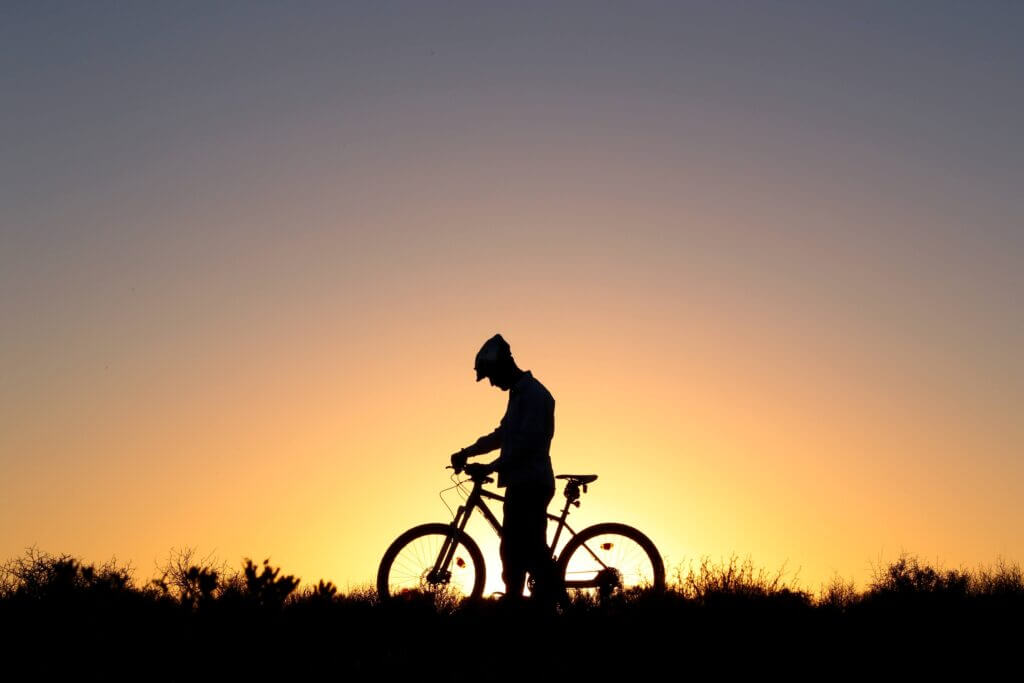 a silhouette of a cyclist stand beside his bicycle with the sun setting behind him