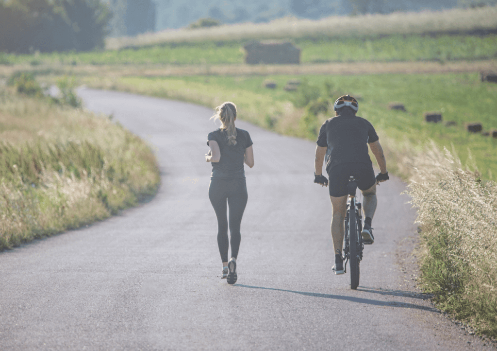 a woman running and a man alongside her cycling on a bike, on a road with fields either side.