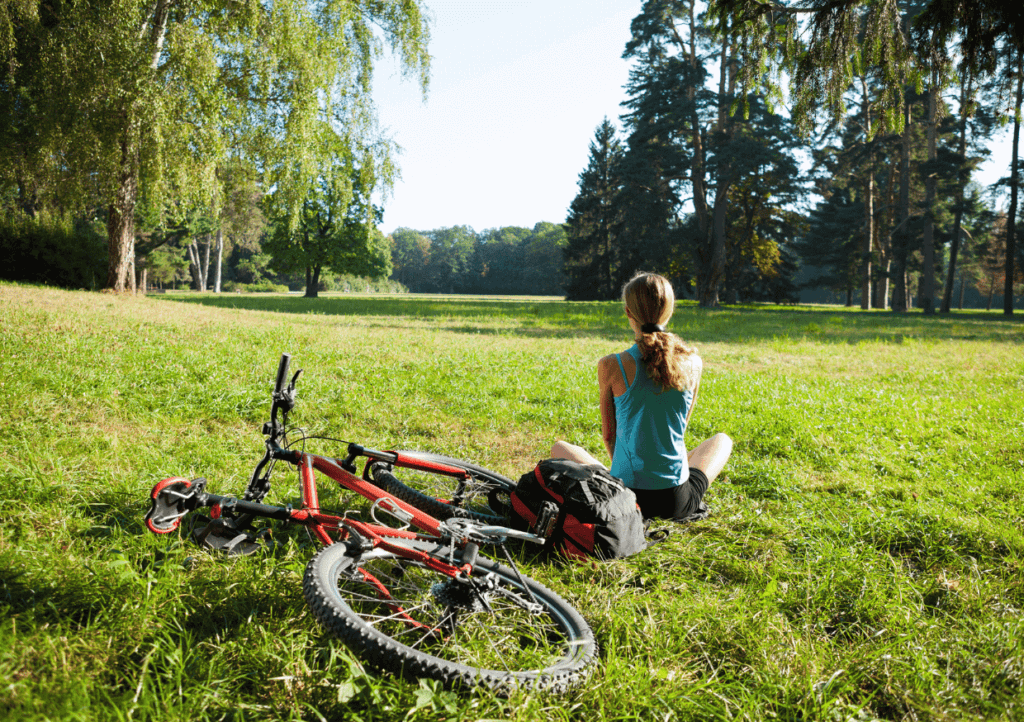 a woman sitting on the grass, beside her bike, in a large field with trees