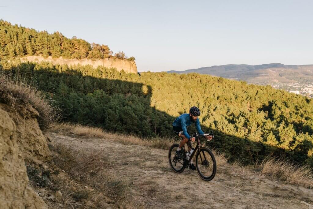 a male cyclist riding a gravel bike up a dirt track with a a forest behind him