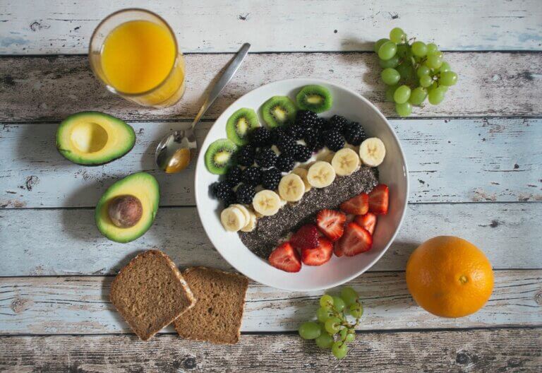 fruit on top of yogurt in a bowl, with brown bread, avocado, and orange juice beside it.