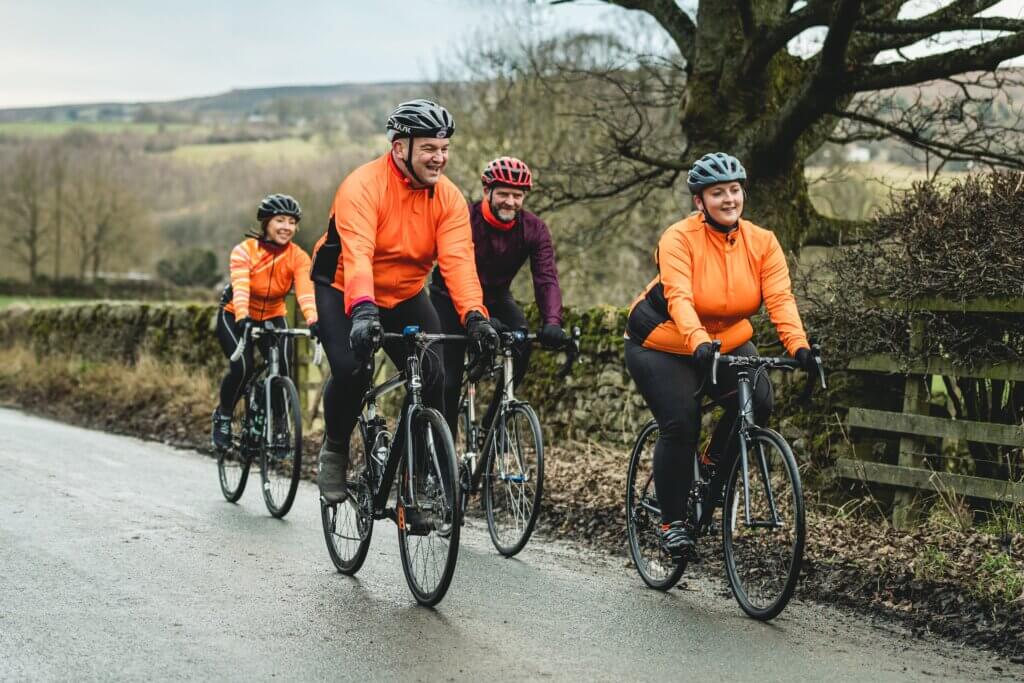 A group of cyclists smiling in maroon and orange jackets riding along a country lane on an Autumnal day