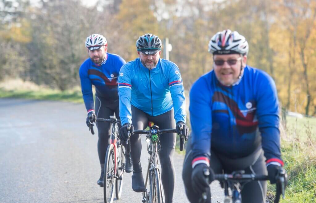 Three male cyclists in blue jackets and jerseys riding their bikes along a country lane in the countryside.