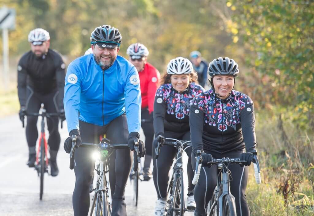 A group of mixed cyclists riding along a country lane smiling