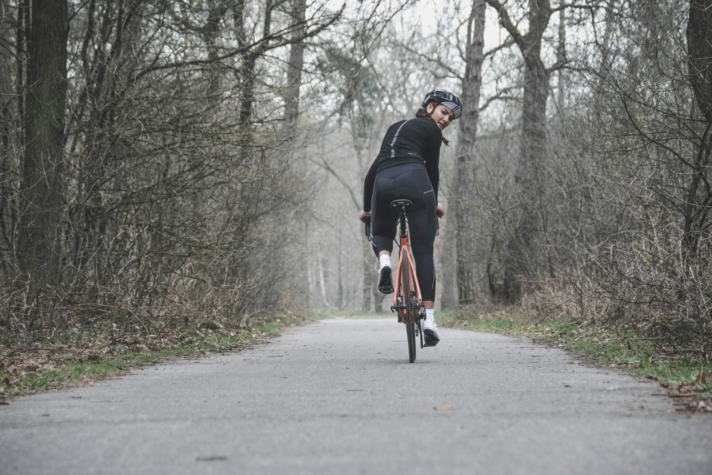 a woman looking over her shoulder as she cyclists down a road through a forest.